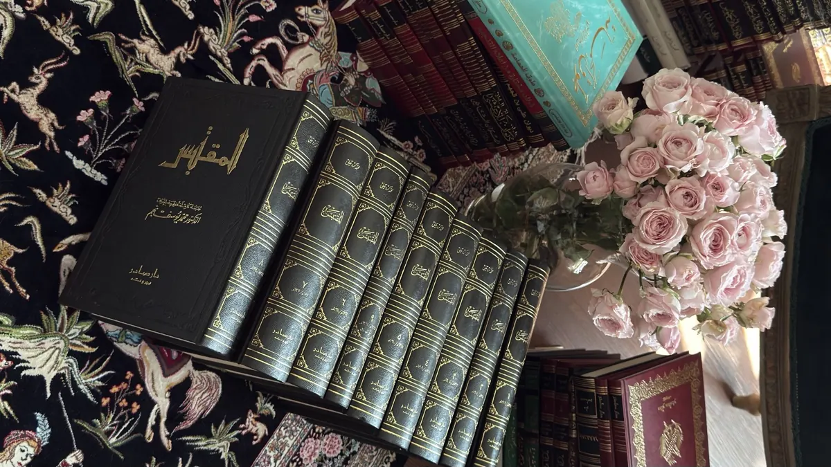 كتاب أسود مذهّب فوق مجموعة مجلّدات فضّية الأكعُب، وباقة ورد وردي على سجادة فارسية سوداء