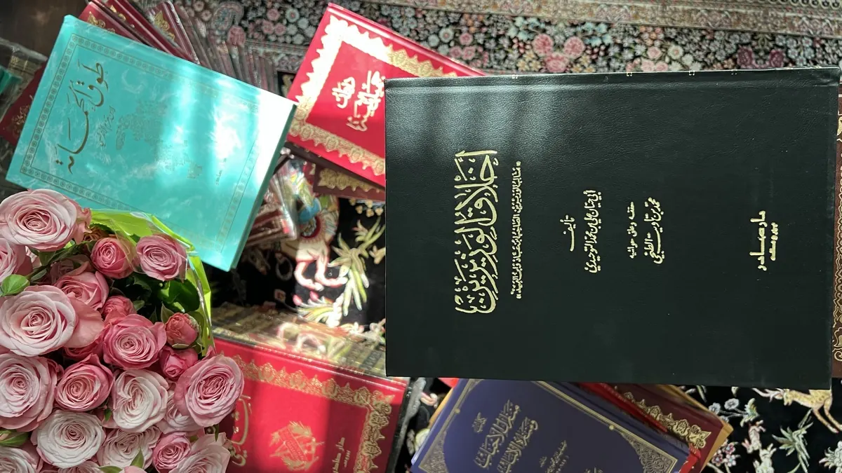 كتاب داكن مذهّب العنوان بجانب كتاب فيروزي وكتب حمراء وباقة ورد وردي